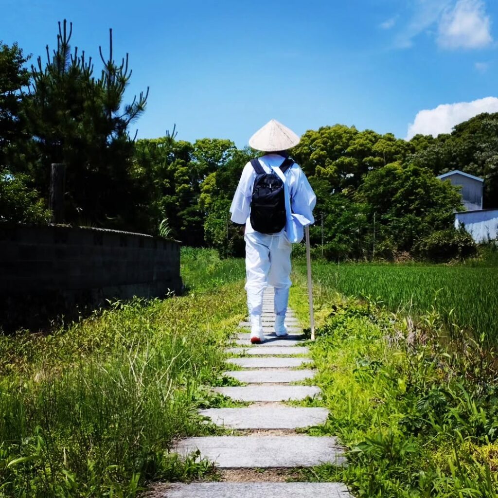 A pilgrim in traditional white clothes walks a grassy trail on the Shikoku Pilgrimage.