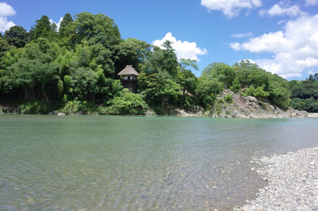 A scenic view of Garyu Sanso in Ozu, Ehime, Japan, looking across the clear Hiji River. The historic thatched-roof sukiya-style villa stands amidst a dense forest, including a prominent bamboo grove, with exposed rocks nearby.