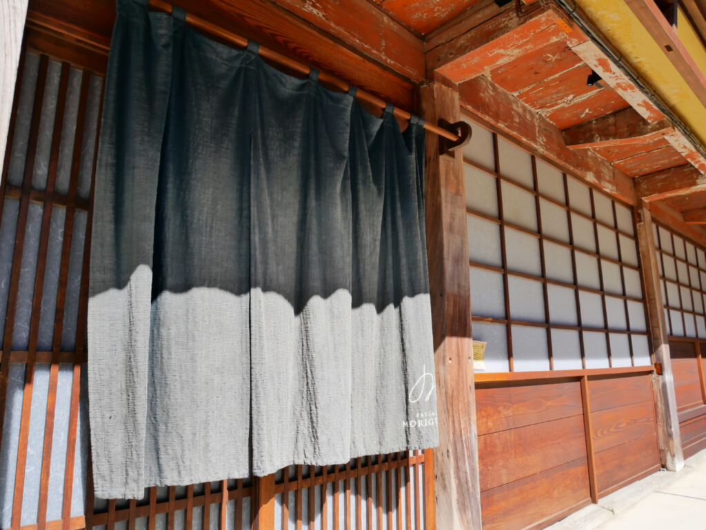 A traditional noren curtain with the logo of PAYSAGE MORIGUCHI hanging at the entrance of the renovated historical boutique hotel.