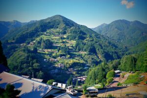 A scenic view of a mountainside village in the Iya Valley, Tokushima, Japan, with traditional houses nestled among dense cedar forests, terraced retaining walls, and winding roads.