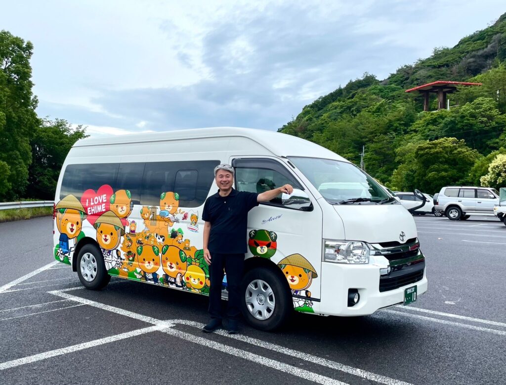 A man and an "I LOVE EHIME" themed Toyota HiAce van featuring the popular Mican mascot in pilgrim and fishing attire, perfect for showcasing local charm during travel in Shikoku.
