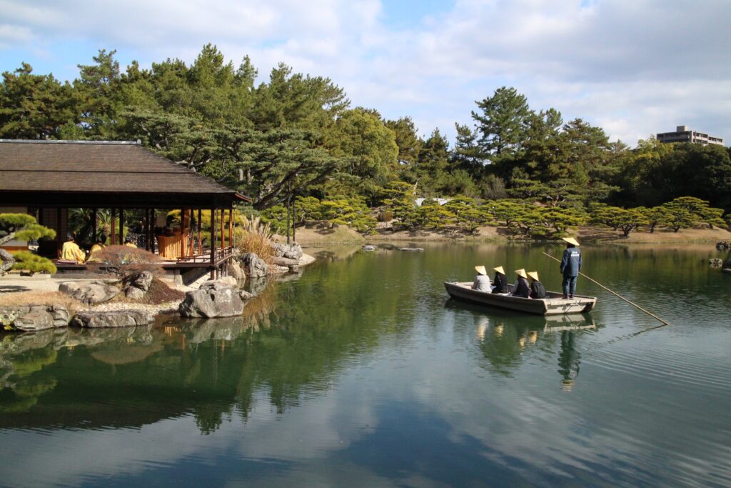 A serene scene in Ritsurin Garden featuring a boatman and passengers in traditional conical hats on a calm pond, reflecting the blue sky and the elegant Kikugetsu-tei pavilion along the rocky shore.