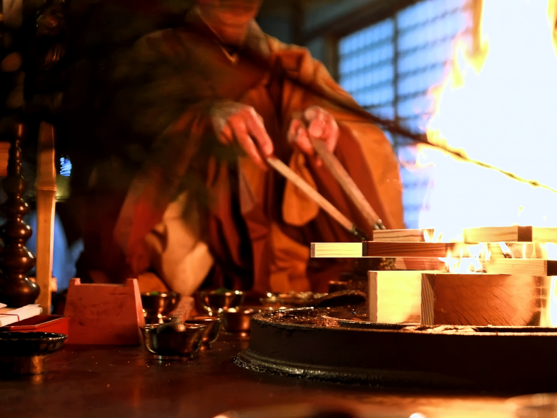A Shingon Buddhist priest performs the goma fire ritual, offering prayers into the sacred flames. The altar is lined with ritual implements used in esoteric Buddhist ceremonies.