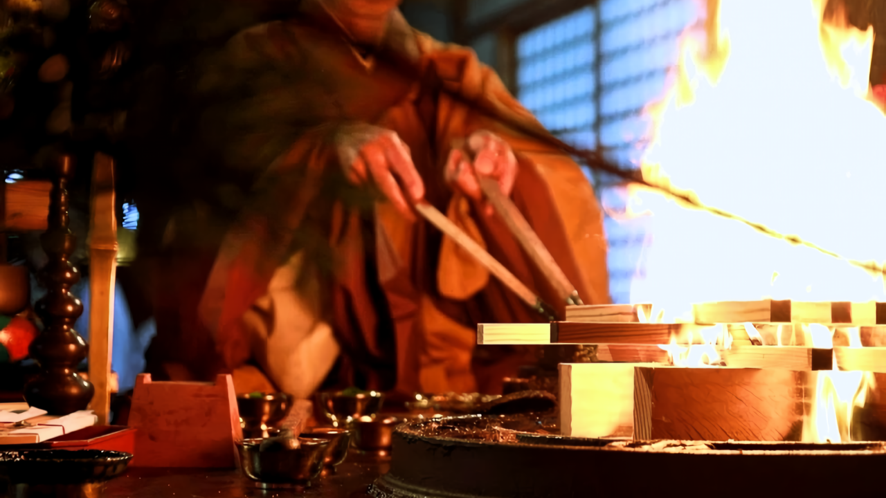 A Shingon Buddhist priest performs the goma fire ritual, offering prayers into the sacred flames. The altar is lined with ritual implements used in esoteric Buddhist ceremonies.