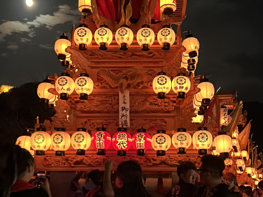 A lantern-covered festival float glows in red and white beneath a hazy moon, with an illuminated castle visible on a distant hilltop.
(22語)