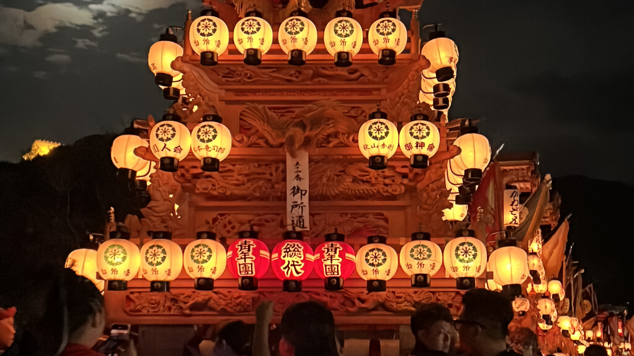 A lantern-covered festival float glows in red and white beneath a hazy moon, with an illuminated castle visible on a distant hilltop.
(22語)