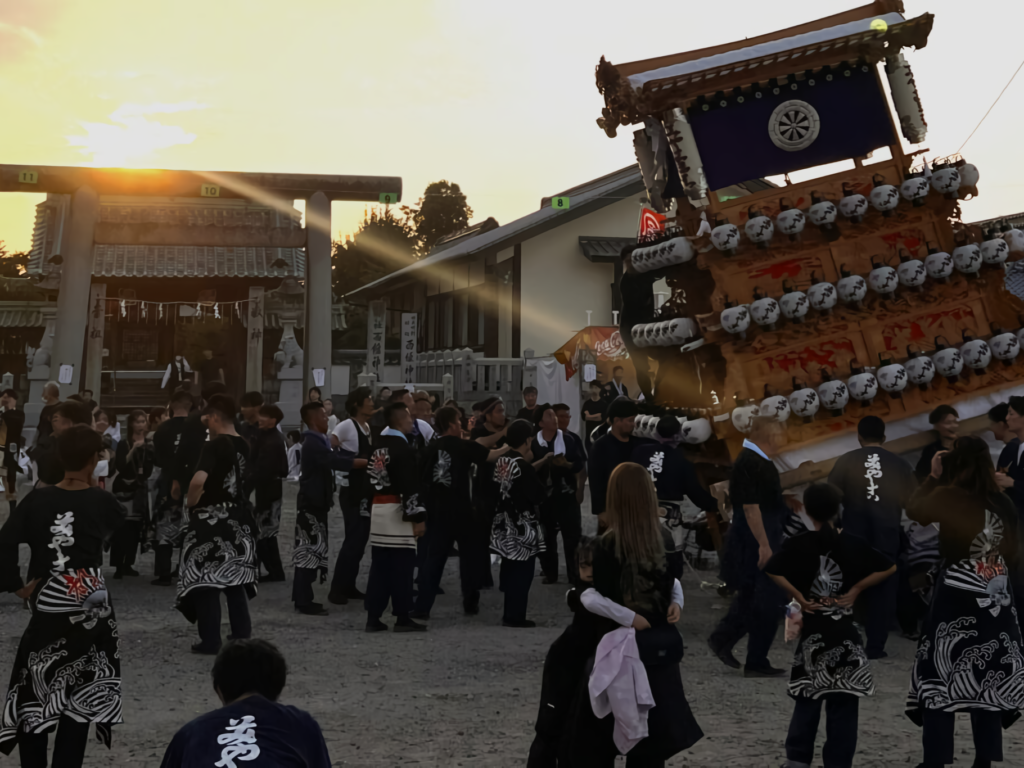 A danjiri float lit with white lanterns is dramatically tilted by festival participants in matching coats before a Shinto shrine torii gate at dusk.