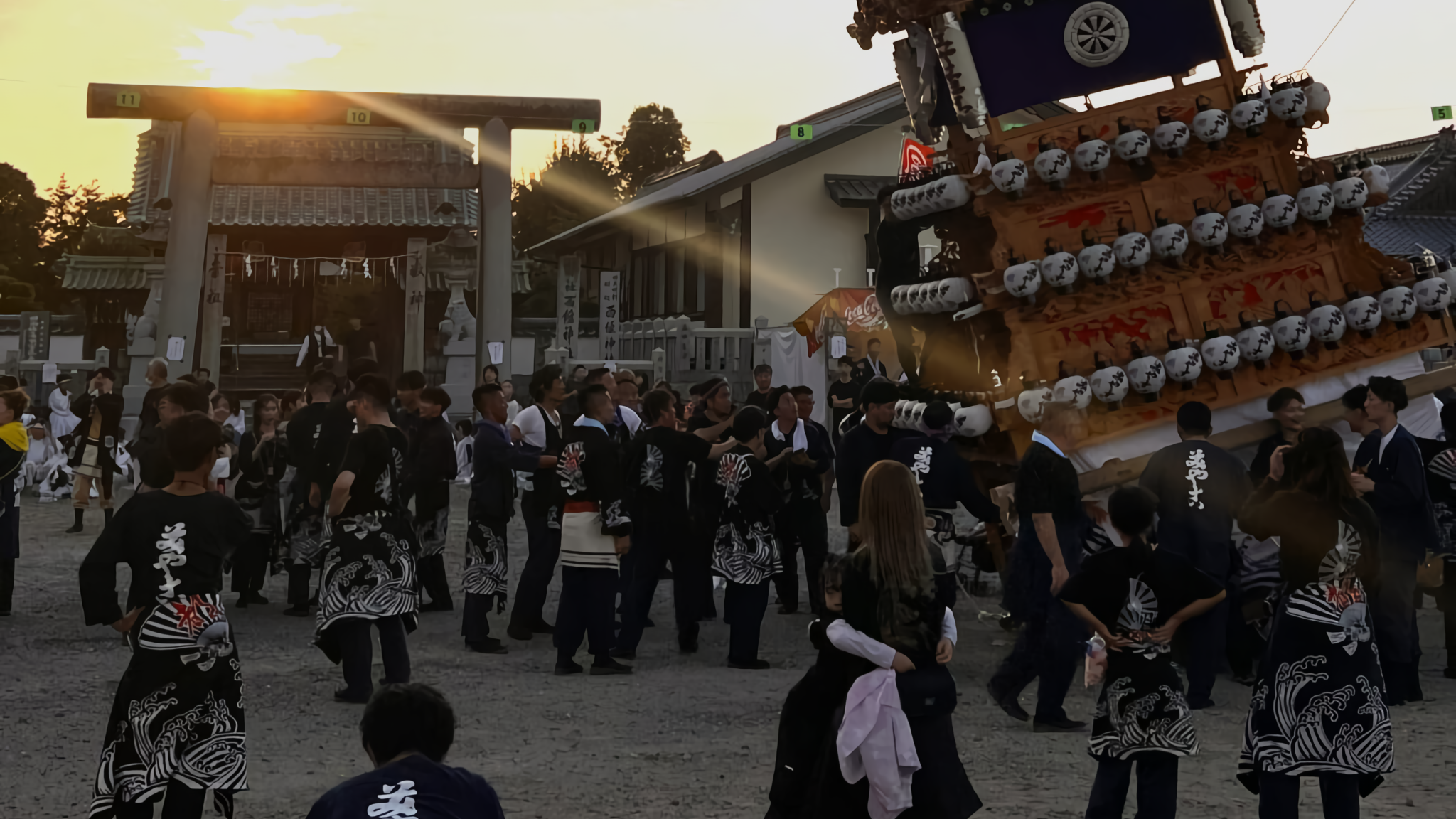 A danjiri float lit with white lanterns is dramatically tilted by festival participants in matching coats before a Shinto shrine torii gate at dusk.