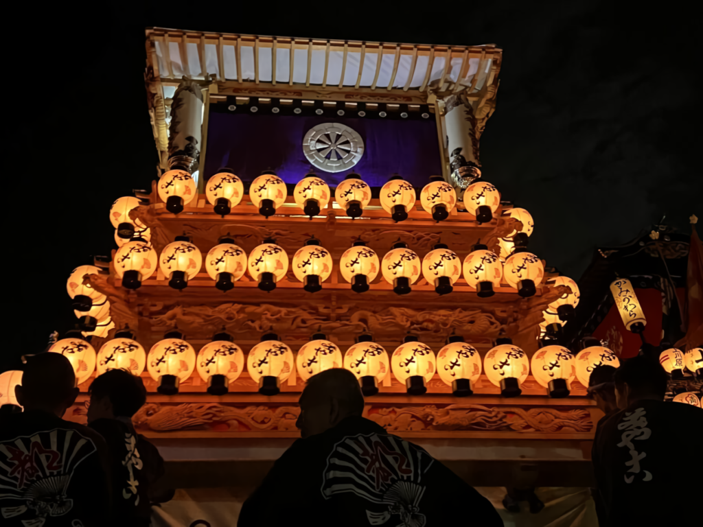 A majestic Japanese festival float glows with white paper lanterns at night, displaying a purple banner with a wheel emblem as participants gather below.