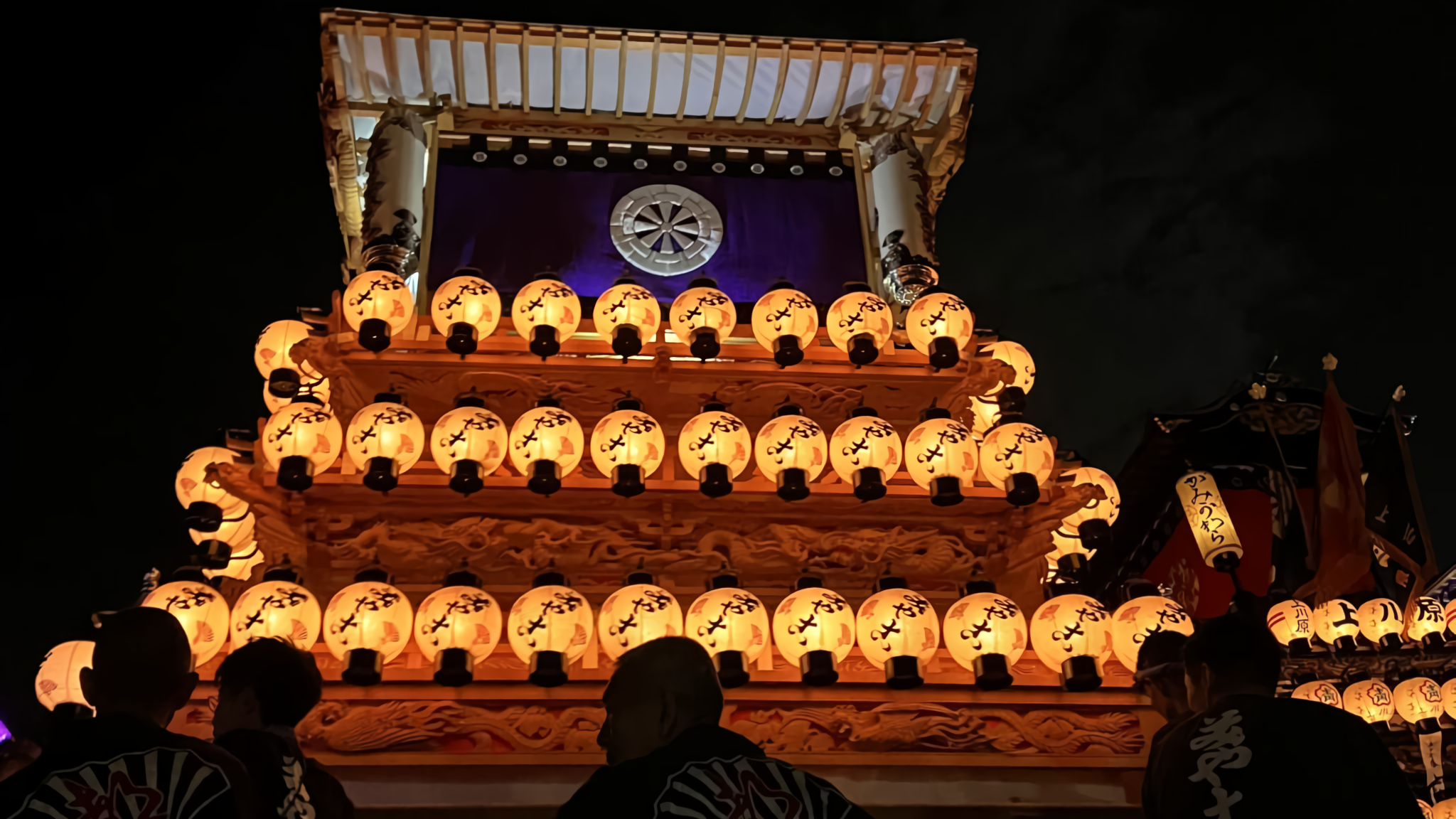 A majestic Japanese festival float glows with white paper lanterns at night, displaying a purple banner with a wheel emblem as participants gather below.
