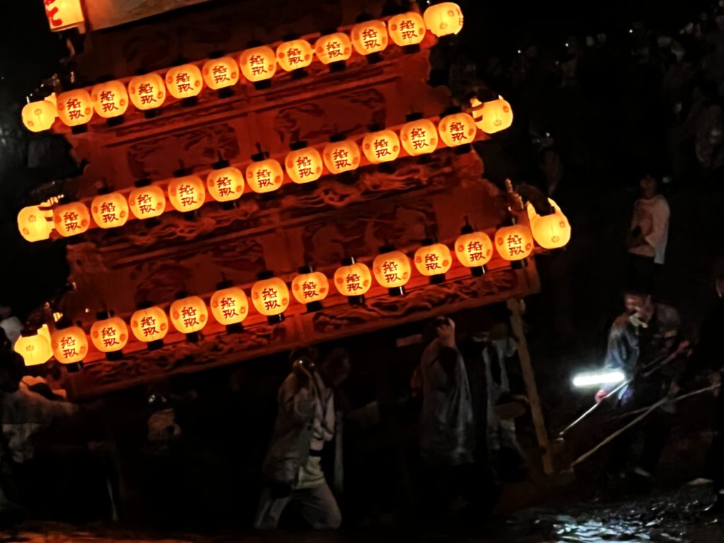 A danjiri float lit with white lanterns is dramatically tilted by festival participants in matching coats before a Shinto shrine torii gate at dusk.