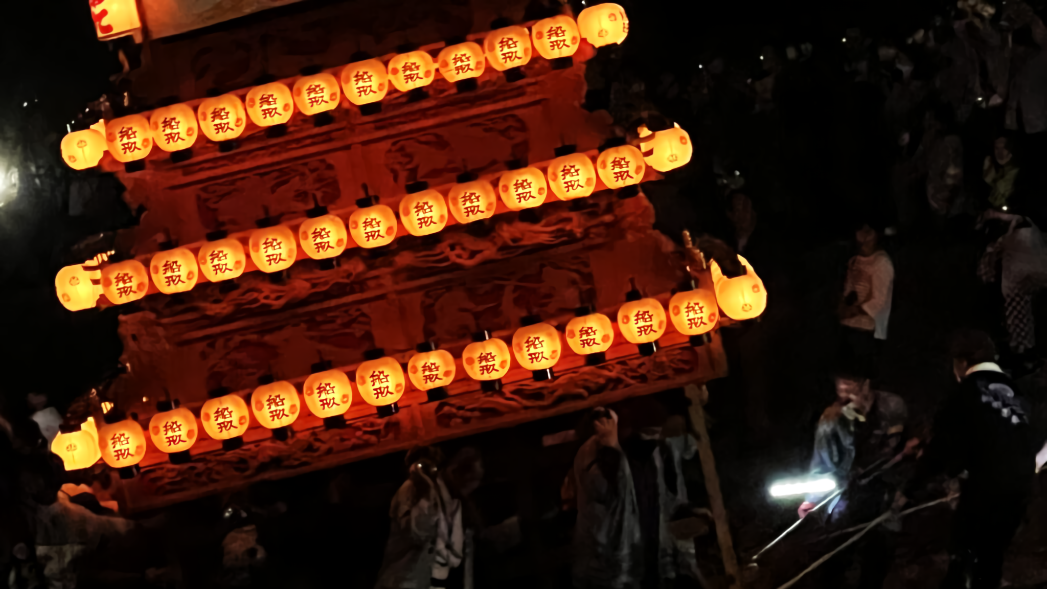 A danjiri float lit with white lanterns is dramatically tilted by festival participants in matching coats before a Shinto shrine torii gate at dusk.