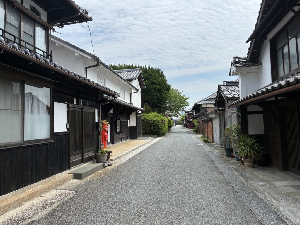 A quiet street in the Unomachi historic district, showcasing traditional white-walled storehouses (kura) and a vintage red Japanese pillar post box.