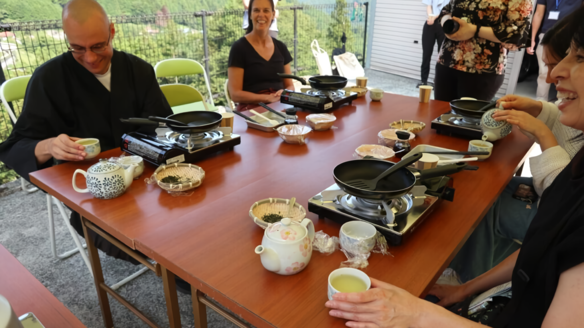 A diverse group enjoys traditional Japanese tea culture on a mountain-view terrace, guided by a monk alongside several women.
