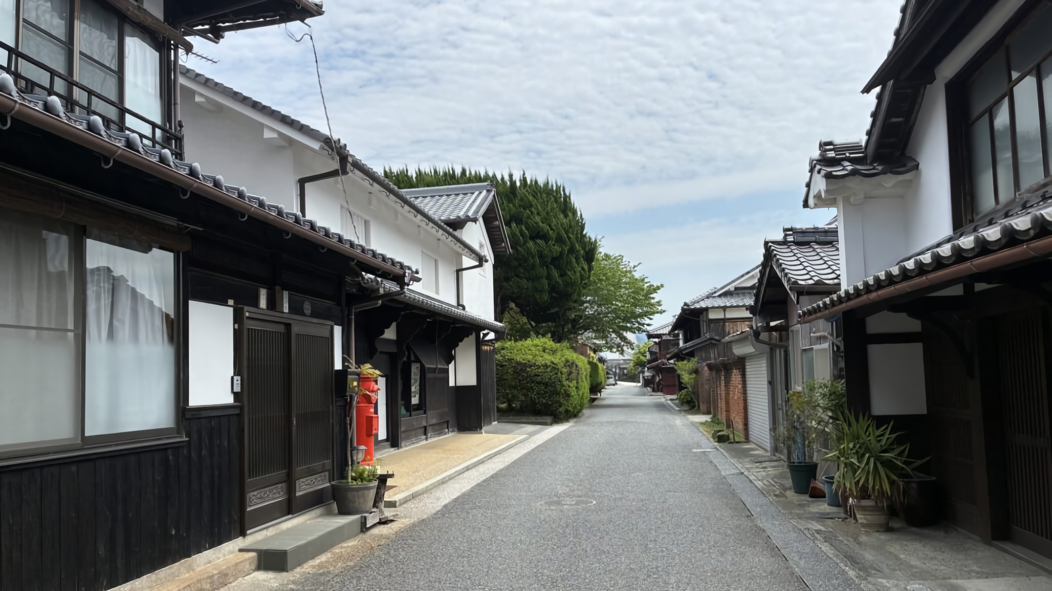A quiet street in the Unomachi historic district, showcasing traditional white-walled storehouses (kura) and a vintage red Japanese pillar post box.