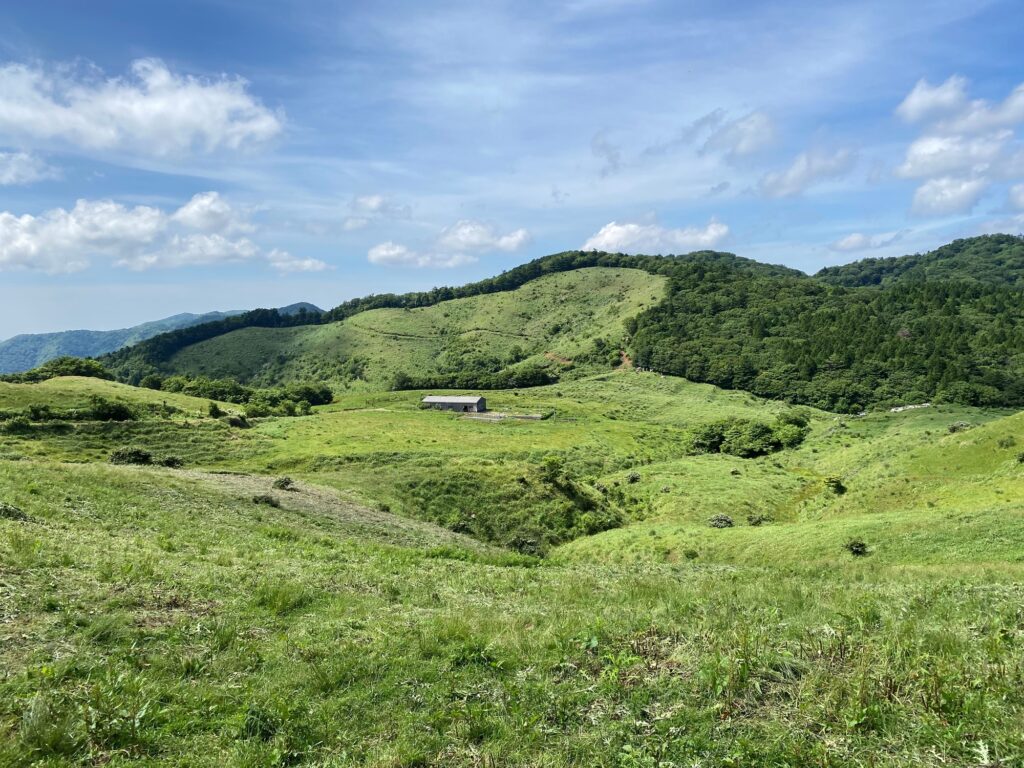 A panoramic view of the lush green rolling hills and meadows of the Onogahara plateau in Seiyo City, featuring a small barn under a blue sky.