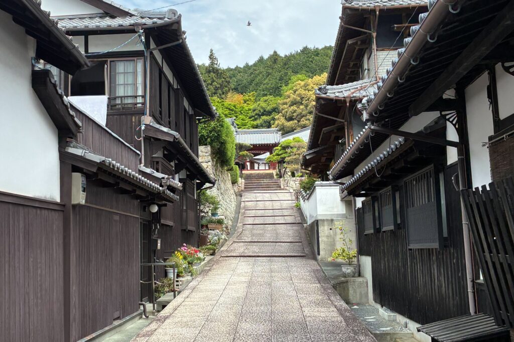 A stone-paved slope lined with traditional Japanese wooden houses in the historic district of Unomachi, leading up to a temple gate nestled in lush green hills.