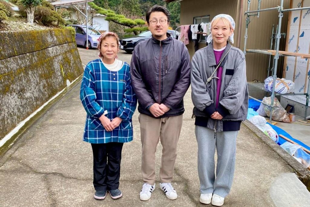 Three people standing together in the scenic Iya Valley region of Japan, with a rural home and mountain greenery in the background.