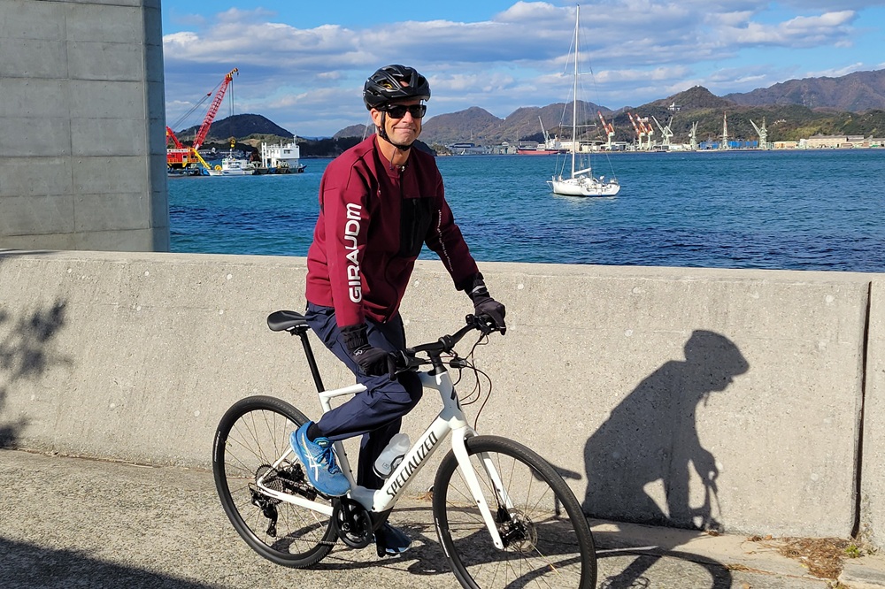 A man cycling along the Shikoku coast on a white Specialized bicycle, wearing a helmet, sunglasses, and a maroon jacket, with the ocean and boats in the background.