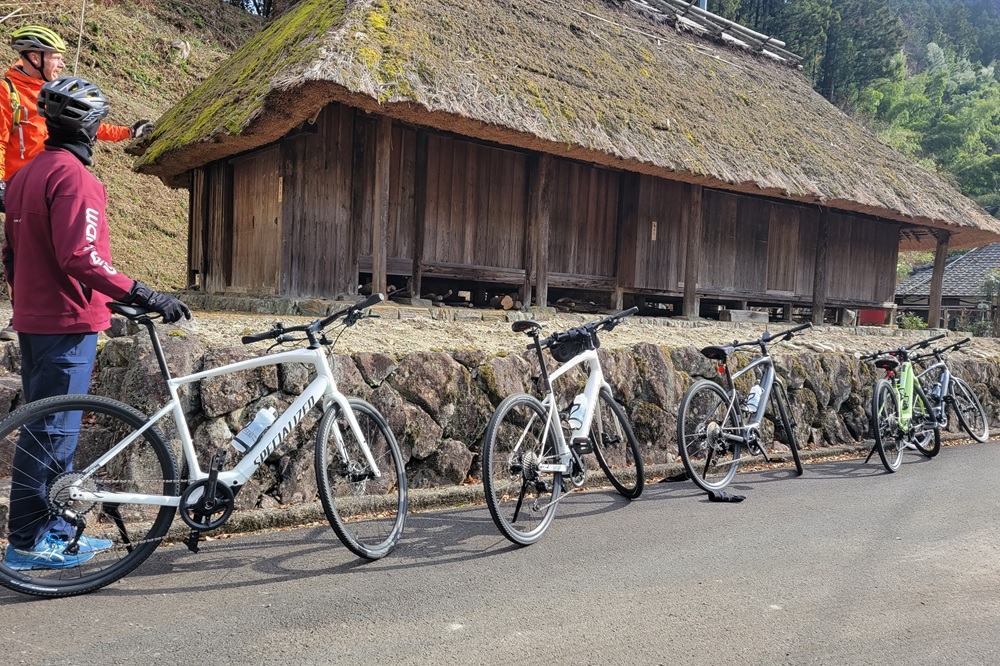 A row of bicycles parked in front of a traditional Japanese thatched-roof house in Shikoku, with two cyclists.
