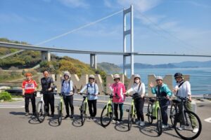 Eight smiling cyclists posing with their Specialized electric bicycles in front of the 'SHIMANAMI' text monument, with the Kurushima Kaikyo Bridge in the background over the Seto Inland Sea