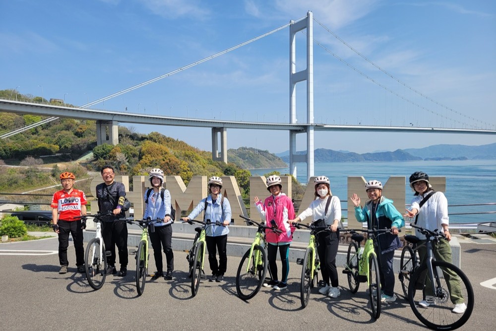 Eight smiling cyclists posing with their Specialized electric bicycles in front of the 'SHIMANAMI' text monument, with the Kurushima Kaikyo Bridge in the background over the Seto Inland Sea