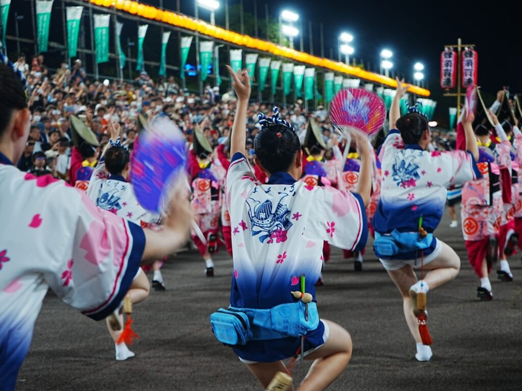 Awa Odori dancers in white and navy yukata performing the energetic "ren" dance step with fans raised at night