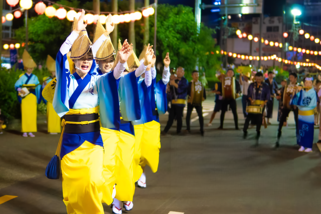Female Awa Odori dancers in blue and yellow costumes dancing with raised hands under festival lanterns at night