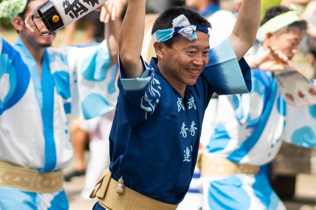A smiling male Awa Odori dancer from the Bushuren group wearing a blue happi coat and dancing with raised arms