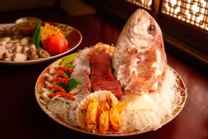 Traditional Japanese sashimi arrangement on a ceramic plate, centered with a whole red seabream head, slices of fresh tuna, and prawns on shredded daikon radish. Another dish with vegetables is visible in the background.