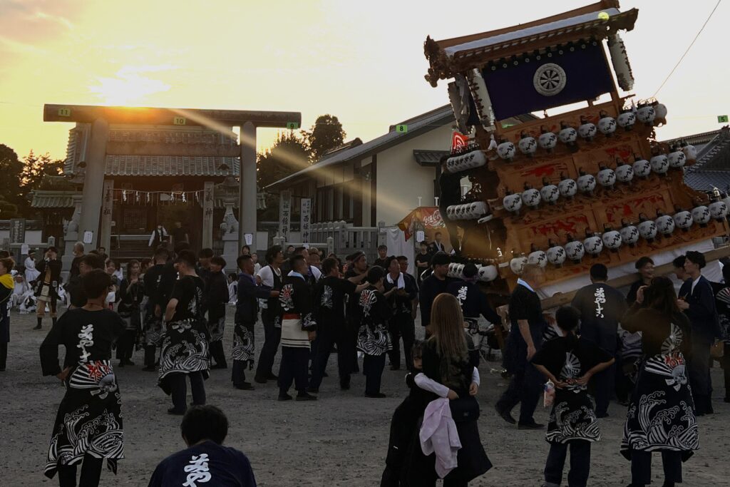 A festival scene at dusk in front of a Shinto shrine torii gate, where a large wooden danjiri float decorated with white lanterns is dynamically tilted by a crowd of participants wearing matching traditional festival coats.