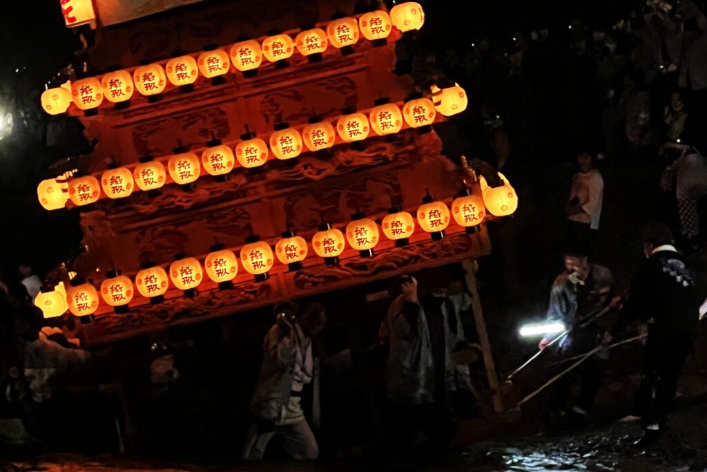 A close-up nighttime shot of a wooden festival float illuminated by rows of glowing orange paper lanterns with Japanese kanji characters, as people carry it through splashing water during a river crossing ritual.