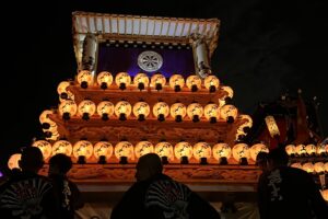 The front view of a majestic Japanese festival float illuminated by numerous white paper lanterns at night, featuring a dark purple banner with a wheel emblem, with the silhouettes of festival participants in the foreground.