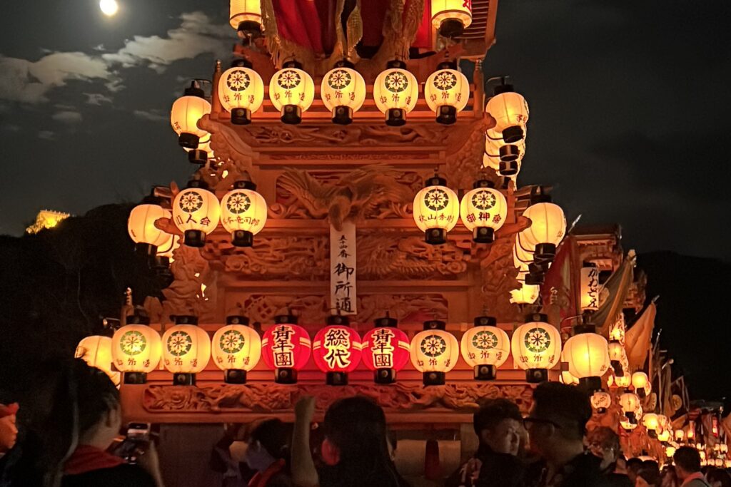 A vibrant night festival scene showing a wooden float heavily decorated with glowing white and red lanterns featuring various Japanese text, set against a night sky with a hazy moon and a illuminated castle visible on a distant hilltop.