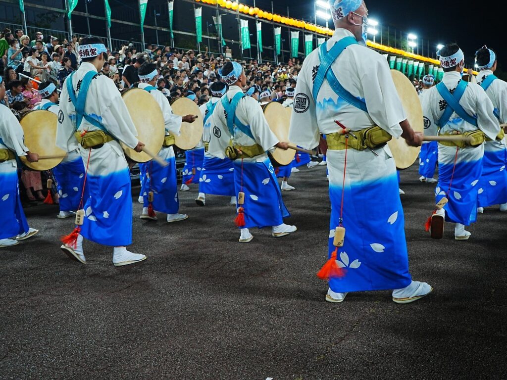Awa Odori drummers in white and blue happi coats striking large taiko drums during a night parade