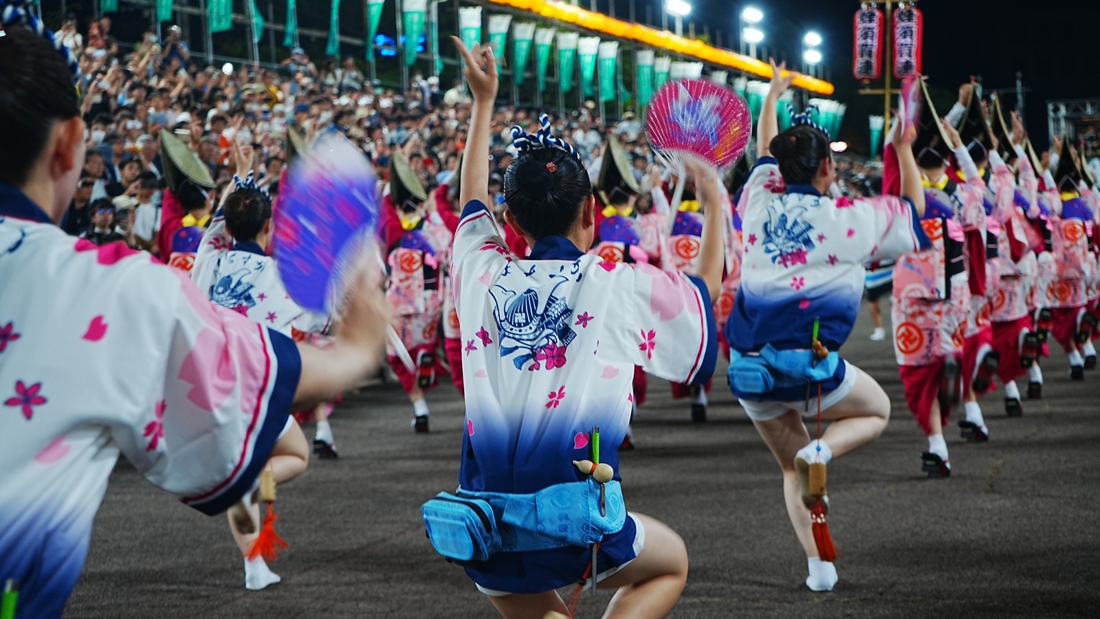 Awa Odori dancers in white and navy yukata performing the energetic "ren" dance step with fans raised at night