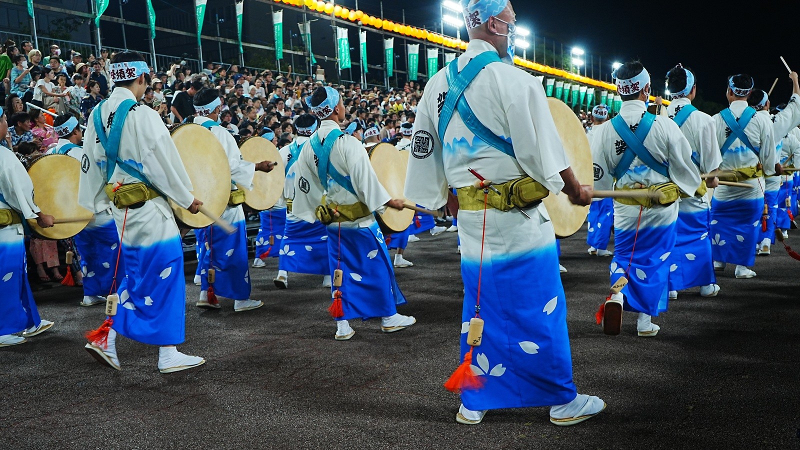 Awa Odori drummers in white and blue happi coats striking large taiko drums during a night parade