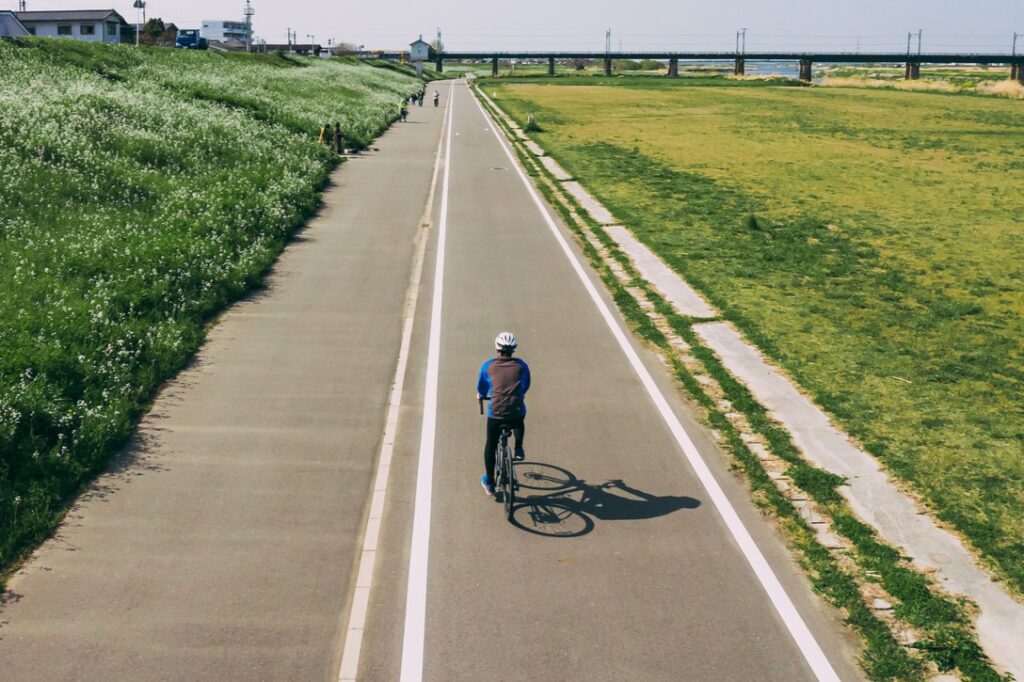 A lone cyclist wearing a helmet is riding a bicycle on the left side of a straight, paved path between a green levee and a large field.
