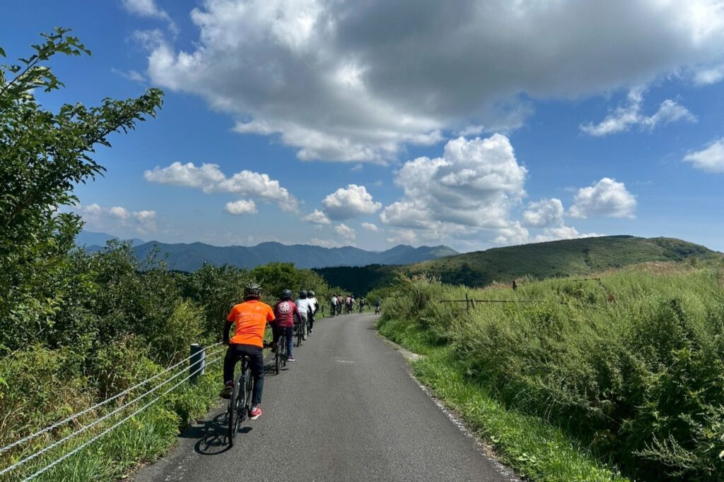 A group of people riding bicycles in a single file line on a paved road through the scenic, grassy hills of the Shikoku Karst under a blue sky with fluffy white clouds.