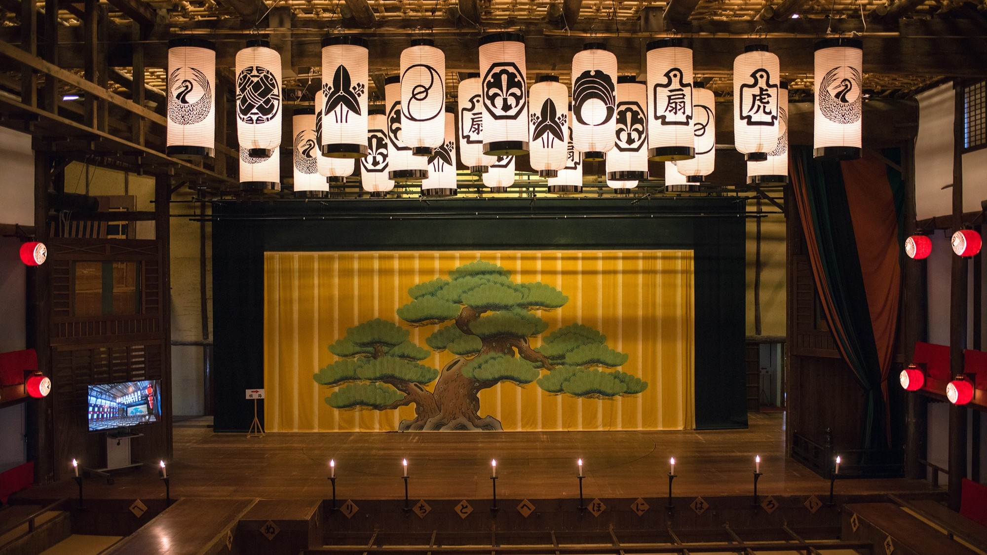 Interior view of Kanamaruza kabuki theater stage with painted pine backdrop and rows of hanging paper lanterns bearing actor crests