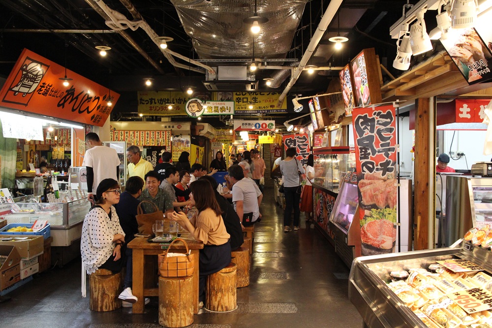 The bustling interior of Hirome Market in Kochi, with people dining at tables surrounded by local food stalls.