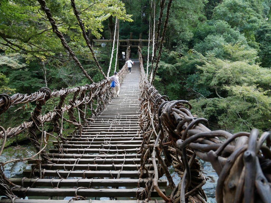 Two visitors cross the Iya Valley Kazura Bridge, its vine-woven walkway swaying above the river, surrounded by dense green forest deep in the Tokushima mountains.