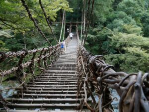 Two visitors cross the Iya Valley Kazura Bridge, its vine-woven walkway swaying above the river, surrounded by dense green forest deep in the Tokushima mountains.