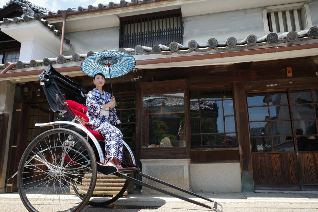 A Japanese woman in an indigo-dyed kimono sits atop a rickshaw in front of a period building in Wakimachi, Tokushima.