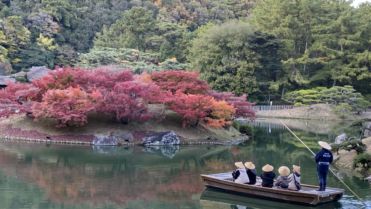 Passengers in traditional straw hats enjoy a boat ride past fiery red and orange maples reflected in still water.