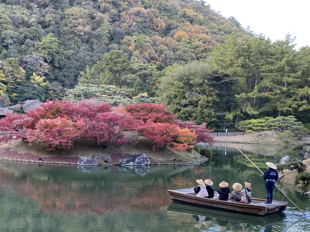 Passengers in traditional straw hats enjoy a boat ride past fiery red and orange maples reflected in still water.