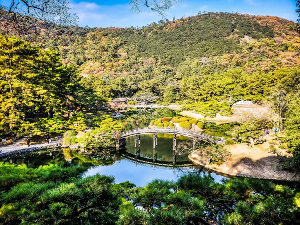 A sweeping panorama showcases the arched bridge, mirror-like pond, sculpted pines, and golden-tinged hillside in warm afternoon light.