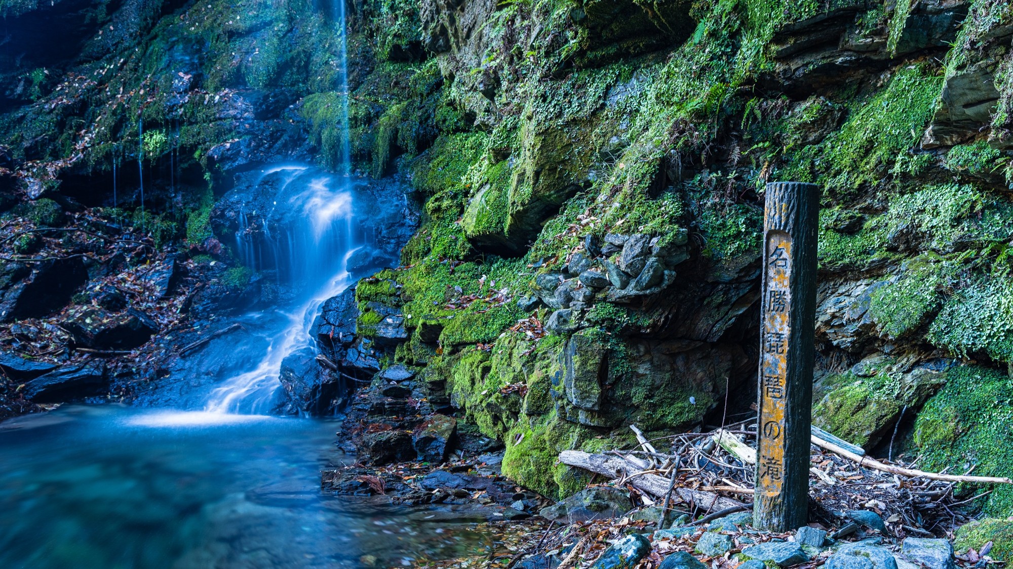 A wooden marker post reading "Biwa Falls" beside the moss-covered rock face of the waterfall