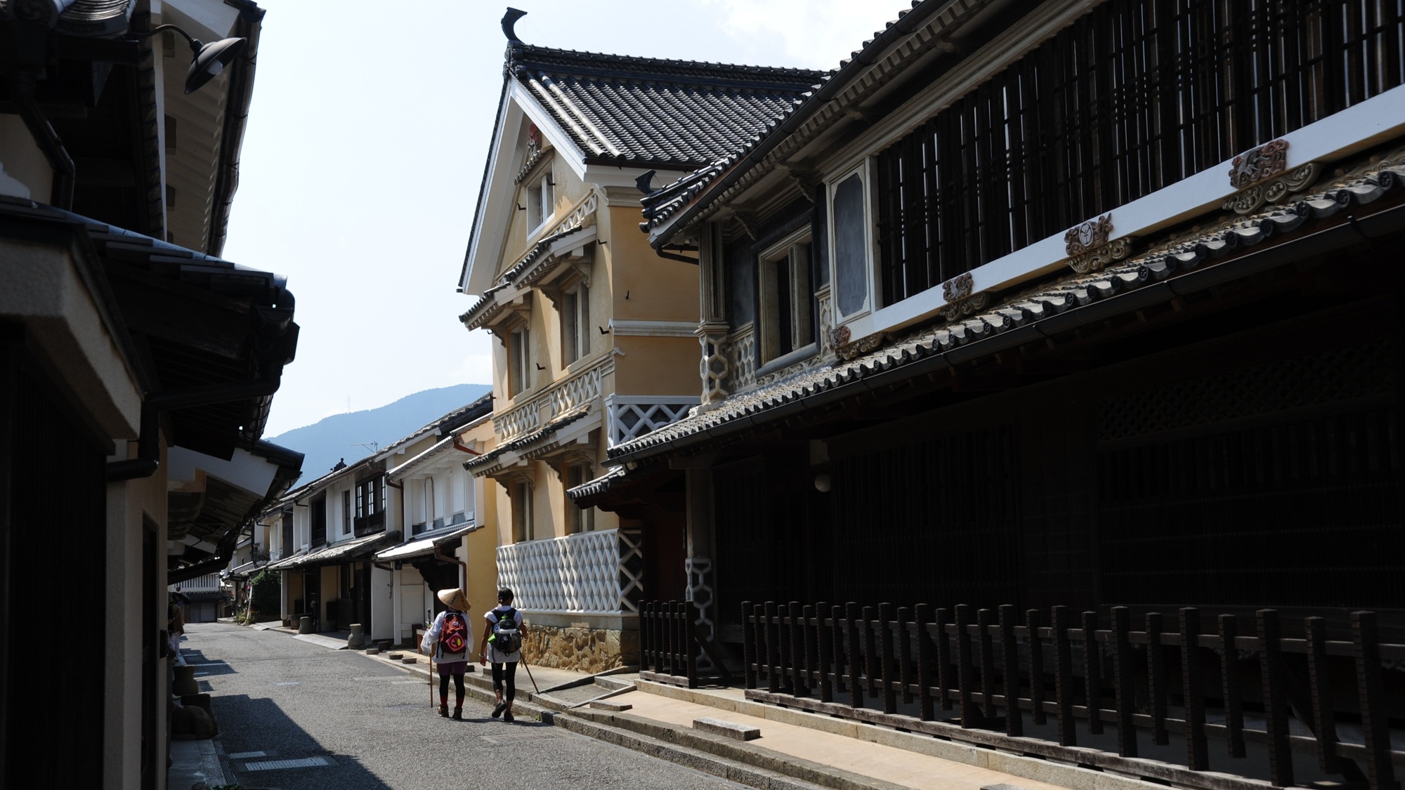 Two travelers in woven sedge hats stroll past ochre plaster merchant houses in Uchiko, where careful restoration keeps the Edo and Meiji streetscape alive.