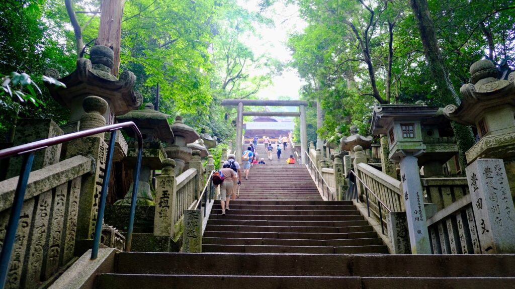 Climbers pause on the steepest stretch, walking sticks in hand, as another torii appears through the summer canopy — proof the top is still coming